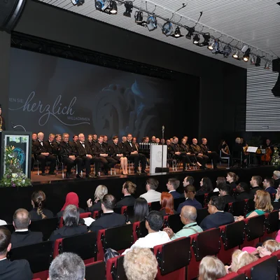 Ein großer Saal bei einer akademischen Feier: Publikum sitzt in Reihen, auf der Bühne eine Gruppe in festlicher Amtstracht, ein Redner am Pult, Beleuchtung von oben; Willkommensprojektion im Hintergrund.