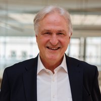 Porträtfoto von Rektor Peter Moser, der lächelnd in die Kamera blickt, gekleidet in einem dunklen Anzug mit weißem Hemd, vor dem Hintergrund des Seminarraums im Studienzentrum.