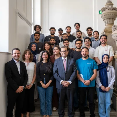 A large group of students and university staff pose on a stone staircase with ornate railings in a historic interior. Several people in formal attire stand at the front, with others in casual clothing behind them.