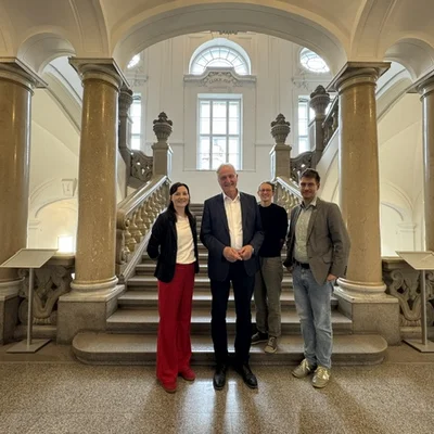 Vier Personen stehen vor einer historischen, säulengerahmten Treppe in einem hellen Foyer; sie blicken in die Kamera.